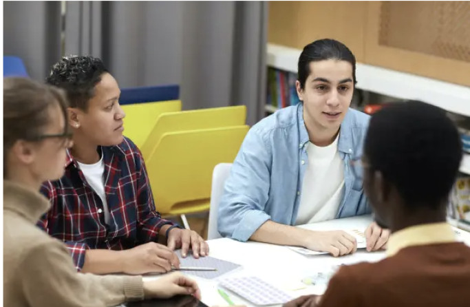 Students sit around a library table while one speaks.